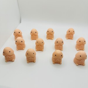 Op de afbeelding: Een groep van 12 kleine, perzikkleurige, squishy speelgoedjes met zwarte ogen. De speelgoedjes zijn gevormd als een menselijk lichaamsdeel en zijn in een cirkel op een wit oppervlak gerangschikt.