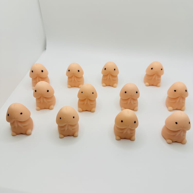 Op de afbeelding: Een groep van 12 kleine, perzikkleurige, squishy speelgoedjes met zwarte ogen. De speelgoedjes zijn gevormd als een menselijk lichaamsdeel en zijn in een cirkel op een wit oppervlak gerangschikt.