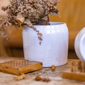 Op de afbeelding: Een witte keramische vaas met een gespikkeld glazuur, gevuld met gedroogde eucalyptus en andere gedroogde bloemen. De vaas staat op een houten tafel met een houten spelbord en spelstukken.