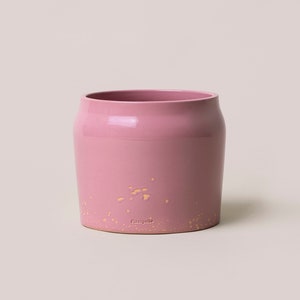 Puede incluir: Un macetero de cerámica rosa con un glaseado dorado moteado. El macetero tiene una forma redondeada y un borde ligeramente acampanado. La parte inferior del macetero está marcada con la palabra "Fiemme"