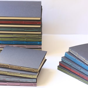 May include: A stack of handmade journals with different coloured covers and stitching. The journals are bound with a variety of colours, including blue, green, red, and gold.
