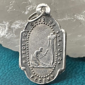 May include: Silver-toned religious pendant with an oval center featuring a kneeling figure and a cross. The pendant has the words "PRECIEUX SANG DE N.S.J.C." and "RECEVEZ" around the edges. A small ring is at the top for attachment.
