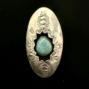 May include: Oval-shaped silver pendant with a turquoise stone center. The pendant features intricate etched designs around the stone and along the edges, with a dark background. The turquoise stone is a light blue-green color.