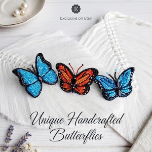 Op de afbeelding: Drie handgemaakte vlinderbroches in blauw- en oranjetinten, met zwarte kralenomtrekken. De vlinders worden getoond op een wit, getextureerd oppervlak. De tekst "Unique Handcrafted Butterflies" staat eronder.