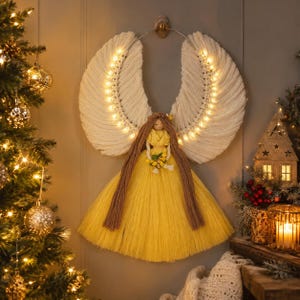 May include: A wall-hanging angel decoration featuring large, white macrame wings illuminated by a string of warm LED lights. The figure has long brown yarn hair, a yellow yarn bodice, and a full, flared yellow skirt, holding a small yellow bouquet.