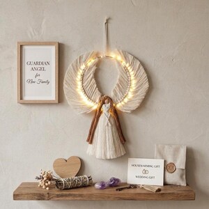 May include: A macrame wall hanging shaped like an angel with illuminated wings, featuring a cream-coloured cord body and hair. Beside it, a framed print reads "GUARDIAN ANGEL for New Family" and a card reads "HOUSEWARMING GIFT WEDDING GIFT".