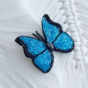 Op de afbeelding: Een vlindervormige broche gemaakt van levendige blauwe glaskralen en omlijnd met zwarte kraaltjes. De vleugels hebben een dicht, getextureerd kralenpatroon, terwijl het centrale lichaam is geaccentueerd met een enkele, donkere, gefacetteerde steen.