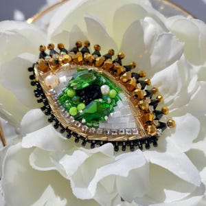 May include: An eye-shaped brooch featuring a central green crystal iris, a black pupil, and white bead accents. The design is bordered by gold and black beads, with gold-toned metallic spikes forming eyelashes along the top edge of the piece.