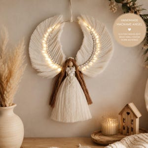 May include: A wall-hanging macrame angel decoration featuring off-white cord wings illuminated by integrated warm LED string lights. The angel has a central figure with brown yarn hair and a long, flowing off-white cord dress suspended from a hoop.