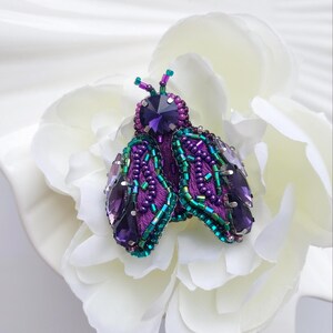 Op de afbeelding: Handgemaakte insectenbroche met paarse, groene en heldere kristallen kralen. De broche heeft een grote, facetgeslepen paarse kristal in het midden, met gedetailleerde vleugels en lichaam. Op een witte achtergrond.