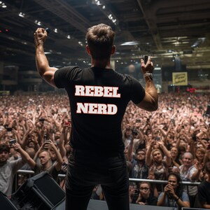 May include: A person wearing a black t-shirt with the text "Rebel Nerd" in white letters with a red outline. The person is standing in front of a large crowd of people.