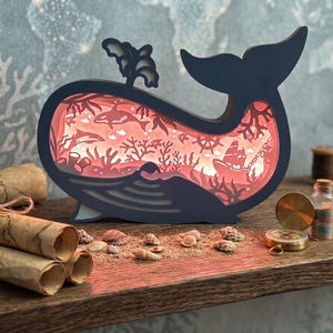 Könnte beinhalten: Eine marineblaue, walförmige Schichtlampe mit einer rot beleuchteten Unterwasserszene. Die Szene umfasst Orcas, Korallen und ein Schiff. Dekorative Gegenstände wie Schriftrollen, Muscheln und ein Kompass sind auf einer Holzoberfläche angeordnet.