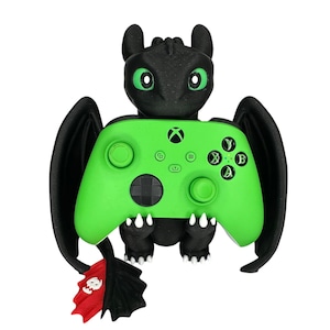 May include: A black dragon-shaped controller holder with a bright green Xbox controller. The dragon has green eyes, black wings, and a red and white emblem. The controller has black buttons and a black directional pad.
