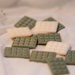 Puede incluir: Un grupo de pequeñas fundidas de cera cuadradas en verde y blanco. Las fundidas de cera están dispuestas sobre una superficie de tela blanca.