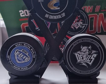 May include: Three black hockey pucks with different logos and text. The top puck is a black and gold puck with the text "OFFICIAL GAME PUCK", "25 YEARS", "IIHF", "2022-23", "GROUPE E", and "IIHF CONTINENTAL CUP". The middle puck is a black and blue puck with the text "2021 ELITE SERIES", "2021", "ELITE SERIES", and "OFFICIAL GAME PUCK". The bottom puck is a black and red puck with the text "ELITE ICE HOCKEY LEAGUE", "DEVILS", and "OFFICIAL WARM-UP PUCK".