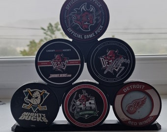 May include: A black hockey puck display stand holding six hockey pucks. The pucks feature team logos and text, including "Mighty Ducks" and "Detroit Red Wings." The top pucks read "Official Game Puck."