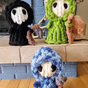 May include: Three finger-knitted Grim Reaper figures in black, green, and blue. Each figure has a white skull face with black eye sockets and holds a brown scythe. The figures are displayed on a stone shelf with the text "Grim Reaper Fingerknit Tutorial".