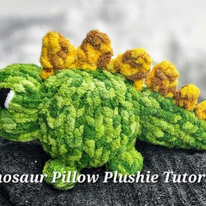 Puede incluir: Un cojín de peluche de dinosaurio de crochet verde y amarillo. El dinosaurio tiene una cola larga y una fila de púas amarillas en la espalda.  Dinosaur Pillow Plushie Tutorial.