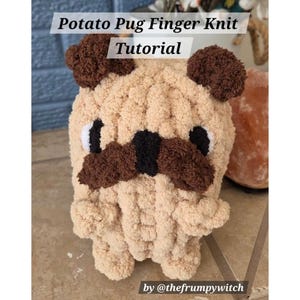 May include: A beige finger-knitted pug dog with brown ears, a brown mustache, and black eyes. The text "Potato Pug Finger Knit Tutorial" is at the top of the image. The text "by @thefrumpywitch" is at the bottom.