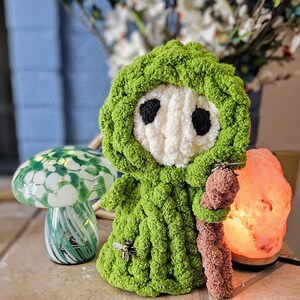 May include: A handmade green crochet doll with a white face and black eyes, wearing a hooded cloak. A small bee charm is attached to the doll's chest. The doll is holding a brown staff. A decorative mushroom lamp and a pink salt lamp are also in the image.