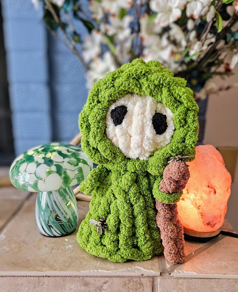 May include: A handmade green crochet doll with a white face and black eyes, wearing a hooded cloak. A small bee charm is attached to the doll's chest. The doll is holding a brown staff. A decorative mushroom lamp and a pink salt lamp are also in the image.