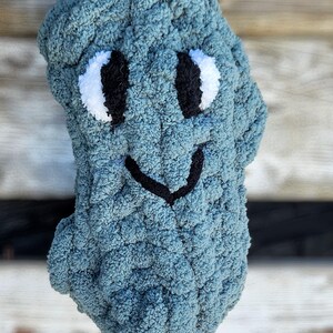 May include: A handmade, blue crocheted plush toy shaped like a friendly creature. It features white and black embroidered eyes and a black stitched smile. The plush is made of a soft, textured yarn.