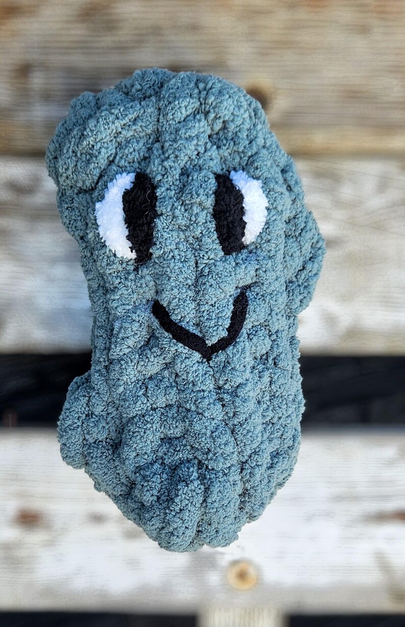 May include: A handmade, blue crocheted plush toy shaped like a friendly creature. It features white and black embroidered eyes and a black stitched smile. The plush is made of a soft, textured yarn.