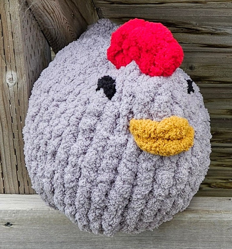 Puede incluir: Un juguete de peluche de pollo de crochet gris con un pomp&oacute;n rojo en la parte superior de la cabeza, un pico amarillo y ojos negros.