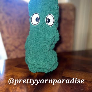 May include: A plush, green monster-like figure with large, embroidered eyes. The figure is made of a soft, textured material and sits on a wooden surface. The text "@prettyyarnparadise" is visible.