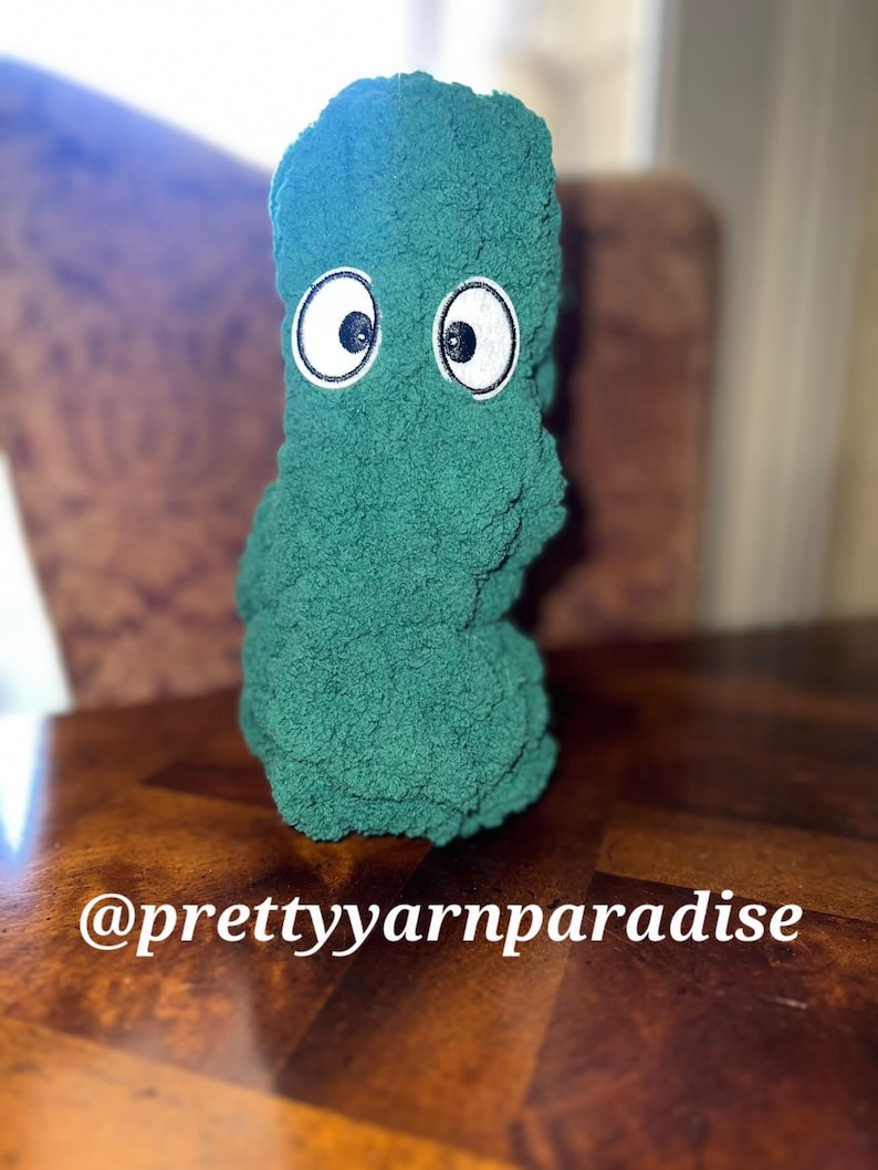 May include: A plush, green monster-like figure with large, embroidered eyes. The figure is made of a soft, textured material and sits on a wooden surface. The text "@prettyyarnparadise" is visible.