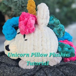 Puede incluir: Un cojín de peluche de unicornio blanco tejido a crochet con una crin y un cuerno de lana azul, rosa y verde. El peluche está sentado sobre una superficie gris. "Unicorn Pillow Plushie Tutorial" está escrito en texto blanco en la imagen.