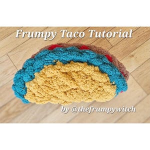 May include: A crocheted taco pillow with a brown, blue, and red shell. The pillow is filled with yellow yarn. The text "Frumpy Taco Tutorial by @thefrumpywitch" is on the wooden surface below the pillow.