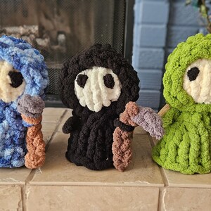 May include: Three handmade crochet figures in blue, black, and green. Each figure has a white face with black eyes, a hood, and holds a staff with a scythe. The figures are displayed on a tiled surface.