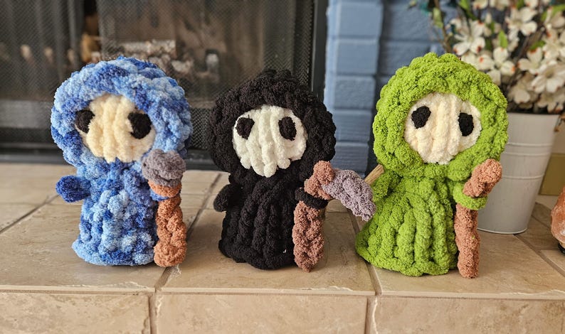 May include: Three handmade crochet figures in blue, black, and green. Each figure has a white face with black eyes, a hood, and holds a staff with a scythe. The figures are displayed on a tiled surface.