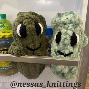 May include: Two handmade crochet creatures in a refrigerator. One is olive green, the other is light blue-green. Both have white and black eyes and a black smiling mouth. The text "@nessas_knittings" is at the bottom.