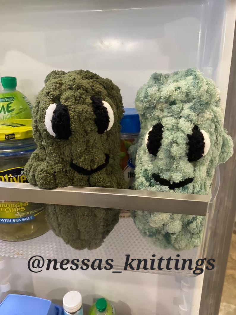 May include: Two handmade crochet creatures in a refrigerator. One is olive green, the other is light blue-green. Both have white and black eyes and a black smiling mouth. The text "@nessas_knittings" is at the bottom.