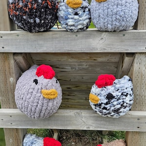 Puede incluir: Seis peluches de pollo tejidos a crochet en varios colores y patrones, incluyendo blanco y negro, gris y beige, dispuestos en un estante de madera.