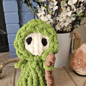 May include: A handmade, green crocheted figure with a white face and black eye details, holding a brown staff. The figure is wearing a hooded cloak. The background includes white flowers in a bucket and a blue brick wall.