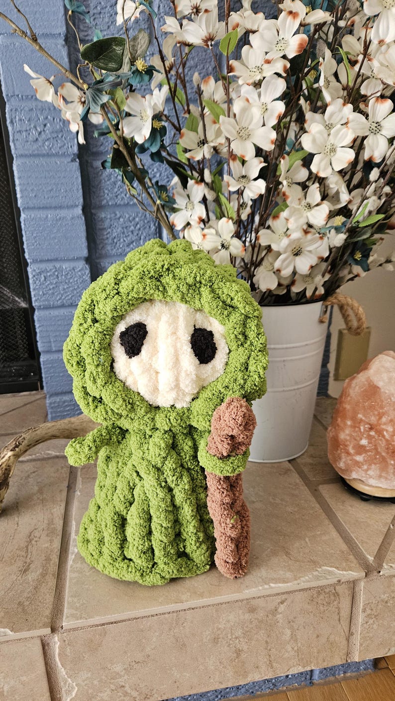 May include: A handmade, green crocheted figure with a white face and black eye details, holding a brown staff. The figure is wearing a hooded cloak. The background includes white flowers in a bucket and a blue brick wall.