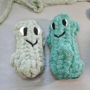 May include: Two plush, handmade monster-shaped toys in shades of gray and teal. Each has a smiling face with black eyes and a black mouth. The toys are made of a soft, textured yarn. A gray yarn is in the background.