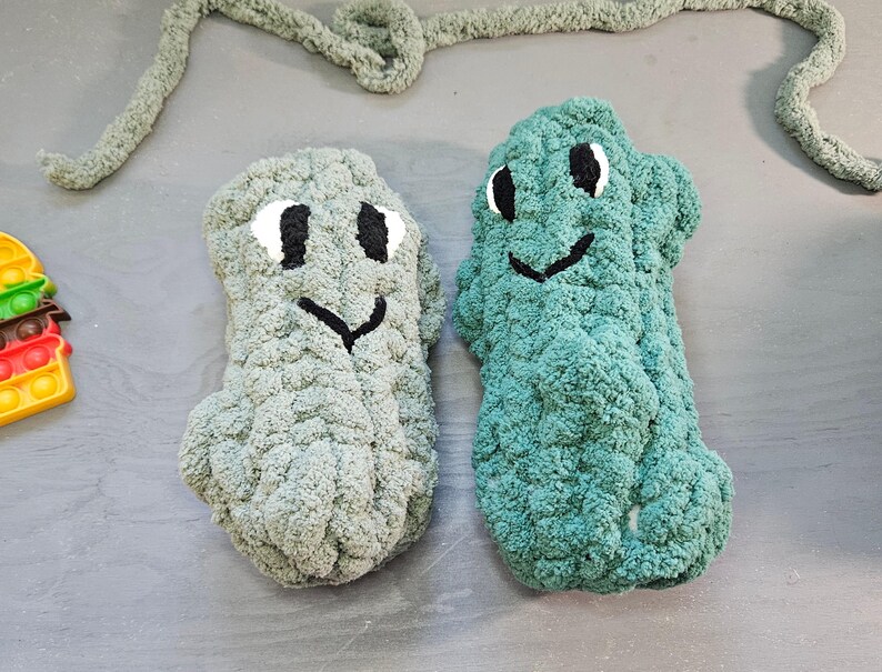 May include: Two plush, handmade monster-shaped toys in shades of gray and teal. Each has a smiling face with black eyes and a black mouth. The toys are made of a soft, textured yarn. A gray yarn is in the background.