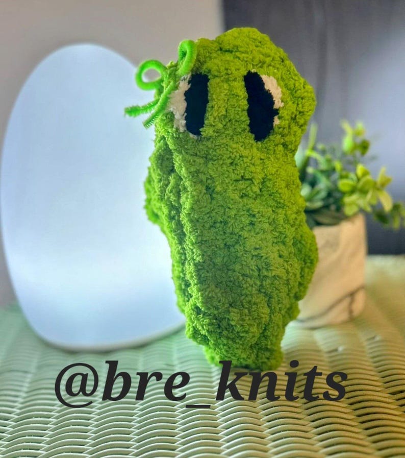 May include: A handmade, green, plush creature with black eyes and a green pipe cleaner accent. The text "@bre_knits" is displayed at the bottom of the image. The plush toy is made of a soft, textured material.