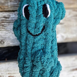 May include: A teal-colored, plush, handmade creature with a smiling face. The eyes are black with white accents, and the mouth is a simple black curve. The soft, textured material gives the toy a cuddly appearance. The background is a weathered wooden surface.