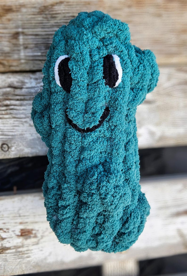 May include: A teal-colored, plush, handmade creature with a smiling face. The eyes are black with white accents, and the mouth is a simple black curve. The soft, textured material gives the toy a cuddly appearance. The background is a weathered wooden surface.