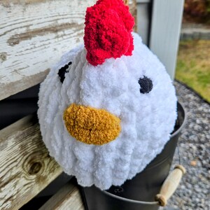 Puede incluir: Pollo blanco de crochet con una cresta de pomp&oacute;n rojo, un pico de hilo amarillo y ojos de hilo negro. El pollo est&aacute; sentado en un cubo negro.
