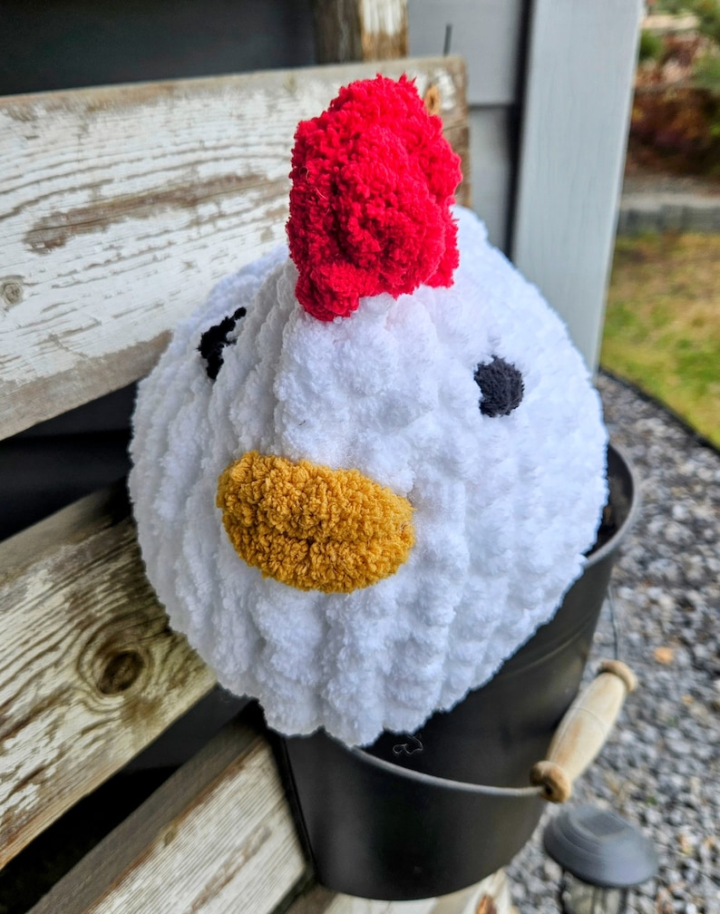 Puede incluir: Pollo blanco de crochet con una cresta de pomp&oacute;n rojo, un pico de hilo amarillo y ojos de hilo negro. El pollo est&aacute; sentado en un cubo negro.