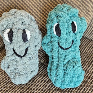 May include: Two handmade, plush, smiling monster-shaped pillows. One is light gray, the other teal. Each has white eyes with black pupils and a black stitched smile. The pillows are made of a soft, textured yarn.