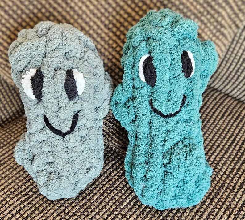 May include: Two handmade, plush, smiling monster-shaped pillows. One is light gray, the other teal. Each has white eyes with black pupils and a black stitched smile. The pillows are made of a soft, textured yarn.