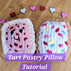 May include: Two plush pillows shaped like a tart pastry. One pillow is pink with red sprinkles and a smiling face. The other pillow is white with colorful sprinkles. The pillows are on a wooden surface with a string of hearts.