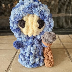May include: A handmade crochet doll in shades of blue and white, with a cream-colored face and dark brown eyes. The doll is holding a brown and gray object, possibly a staff or a toy. The doll is standing on a tile floor.